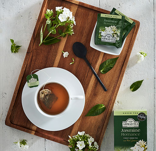 Picture is of a tray containing Jasmine flowers, tea leaves, tea spoon, cup and saucer with tea and a tea bag and tea bags on a small saucer